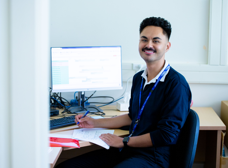 A Health New Zealand office worker sitting at a desk with a computer, holding a pen and paper.