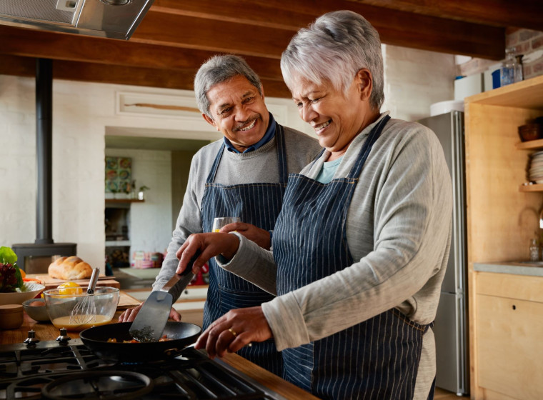 An older man and a woman are smiling as they cook dinner together. 